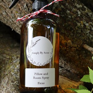 Pode incluir: Uma garrafa de vidro marrom com um bico de pulveriza&ccedil;&atilde;o preto e um r&oacute;tulo branco. O r&oacute;tulo diz "Simply By Anne" em um c&iacute;rculo com um design floral e "Pillow and Room Spray Focus".