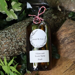 Pode incluir: Uma garrafa de vidro marrom com um bico de pulveriza&ccedil;&atilde;o preto e um r&oacute;tulo branco que diz "Simply By Anne" e "Pillow and Room Spray Fresh". A garrafa est&aacute; amarrada com um cord&atilde;o vermelho e branco.