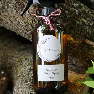 Pode incluir: Uma garrafa de vidro marrom com um bico de pulveriza&ccedil;&atilde;o preto e um r&oacute;tulo branco que diz "Simply By Anne" e "Pillow and Room Spray Sleep". A garrafa est&aacute; amarrada com um barbante vermelho e branco.