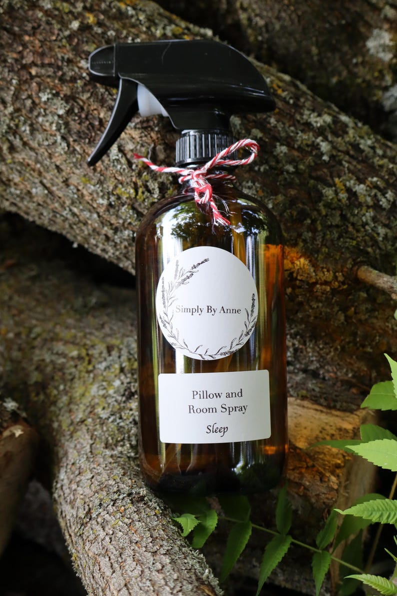 Pode incluir: Uma garrafa de vidro marrom com um bico de pulveriza&ccedil;&atilde;o preto e um r&oacute;tulo branco que diz "Simply By Anne" e "Pillow and Room Spray Sleep". A garrafa est&aacute; amarrada com um barbante vermelho e branco.
