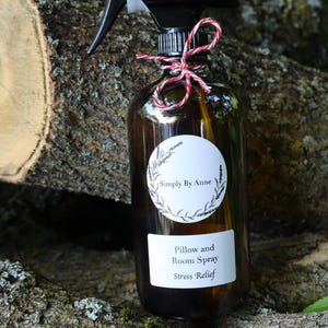 Pode incluir: Uma garrafa de vidro marrom com um bico de pulveriza&ccedil;&atilde;o preto e um r&oacute;tulo branco que diz "Simply By Anne Pillow and Room Spray Stress Relief". A garrafa est&aacute; amarrada com um barbante vermelho e branco e est&aacute; sentada em um peda&ccedil;o de casca de &aacute;rvore.