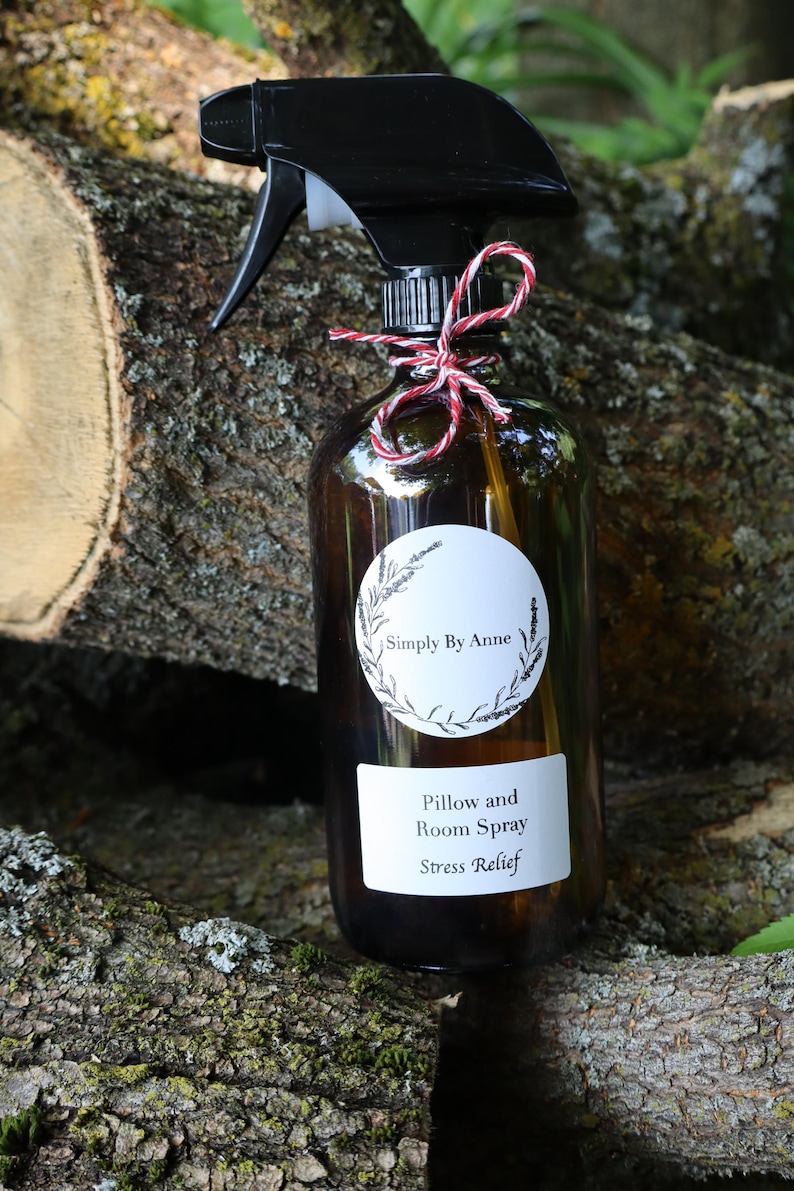Pode incluir: Uma garrafa de vidro marrom com um bico de pulveriza&ccedil;&atilde;o preto e um r&oacute;tulo branco que diz "Simply By Anne Pillow and Room Spray Stress Relief". A garrafa est&aacute; amarrada com um barbante vermelho e branco e est&aacute; sentada em um peda&ccedil;o de casca de &aacute;rvore.