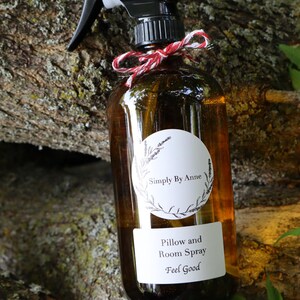 Pode incluir: Uma garrafa de vidro marrom com um bico de pulveriza&ccedil;&atilde;o preto e um r&oacute;tulo branco. O r&oacute;tulo diz "Simply By Anne" em um c&iacute;rculo com um design floral e "Pillow and Room Spray Feel Good" abaixo. A garrafa est&aacute; amarrada com uma fita vermelha e branca.