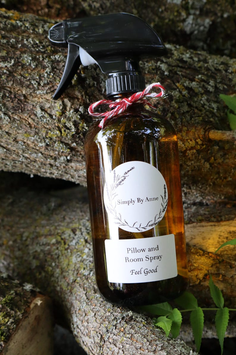Pode incluir: Uma garrafa de vidro marrom com um bico de pulveriza&ccedil;&atilde;o preto e um r&oacute;tulo branco. O r&oacute;tulo diz "Simply By Anne" em um c&iacute;rculo com um design floral e "Pillow and Room Spray Feel Good" abaixo. A garrafa est&aacute; amarrada com uma fita vermelha e branca.