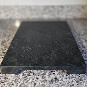 May include: A rectangular, dark gray marble cutting board with a textured surface. The board has a small, rectangular cutout on one side. The cutting board is displayed on a speckled countertop.