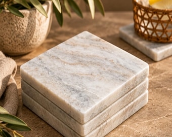 May include: A stack of three square marble coasters featuring a polished surface with natural grey and beige veining. Each coaster has a smooth, flat top and straight edges, showcasing the unique stone patterns of the white marble material.