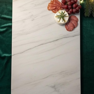 May include: A rectangular white marble serving board with gray veining, holding a selection of gourmet food items. Includes prosciutto, grapes, cheese, and salami. The board rests on a green velvet surface, suggesting a festive or elegant presentation.