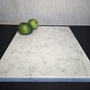 May include: A rectangular white marble cutting board with gray veining. Two green limes rest on the board's surface. The board is set against a neutral background, suggesting a kitchen or food preparation setting.