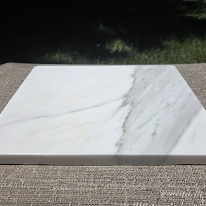May include: A square white marble slab featuring natural grey veining patterns throughout the surface. The stone board has a smooth, polished finish with straight edges and a flat top, showcasing the unique mineral variations of the natural material.