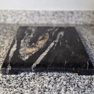 May include: A rectangular cutting board made of black granite with white and gold veining. The board has a smooth surface and small feet on the bottom. This kitchen item is ideal for food preparation.