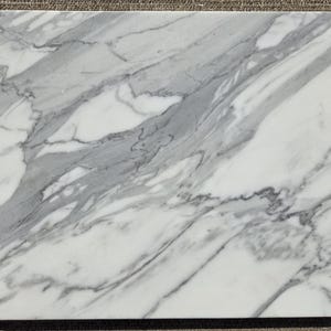 May include: A rectangular marble board featuring a polished white surface with prominent grey veining patterns that flow diagonally across the stone. The edges are smooth and straight, showcasing the natural mineral variations of the material.