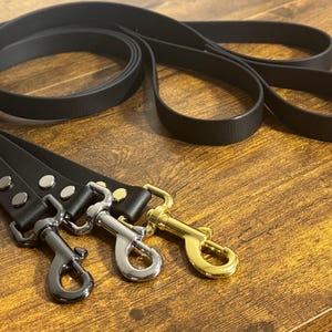 May include: A set of three black leashes featuring smooth, matte-finish straps and metal hardware. Each leash is equipped with a swivel snap hook in a different metallic finish: one in matte black, one in polished silver, and one in gold.