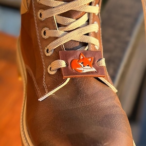 Könnte beinhalten: Brauner Lederstiefel mit beigefarbenen Schnürsenkeln und einem fuchsförmigen Anhänger. Der Stiefel hat eine robuste Sohle und eine hellfarbene Zierleiste. Der Fuchs-Anhänger ist orange und weiß.