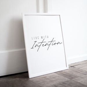 Può includere: Stampa incorniciata bianca con il testo "Live with Intention" in corsivo nero.