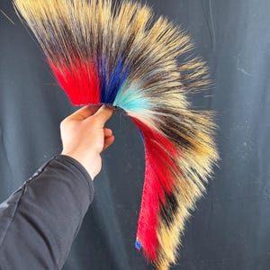 Op de afbeelding: Een decoratief, waaiervormig item gemaakt van bont, met secties geverfd in rood, blauw en bruin. Het bont heeft zwarte accenten. Het item wordt tegen een donkere achtergrond gehouden.
