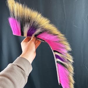 Puede incluir: Un accesorio de piel sintética colorido con un esquema de color rosa, negro, morado y beige. El artículo se sostiene contra un fondo oscuro. El accesorio parece ser una cola o una pieza decorativa para un disfraz o atuendo.