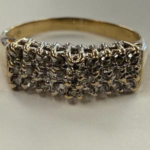 Puede incluir: Un anillo de oro con un engaste rectangular en la parte superior. El engaste está compuesto por varias piedras pequeñas, claras y de corte redondo, dispuestas en tres filas horizontales y sujetas por garras de oro sobre una banda de oro pulida.