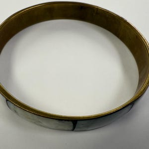 May include: A brass bangle bracelet with a decorative inlay of white and black segments. The bracelet has a circular shape and a polished finish, suitable for everyday wear. The jewellery is displayed on a white surface.