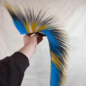 Peut inclure: Une coiffe en fausse fourrure multicolore vibrante. Le design présente une base de fausse fourrure bleue, jaune, noire et crème. La coiffe est tenue par une main sur un fond blanc.