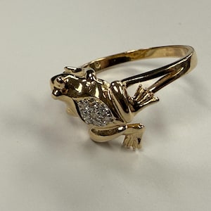Op de afbeelding: Een goudkleurige ring met een kikkerontwerp. De kikker heeft een cluster van kleine, heldere stenen op zijn lichaam. De ring heeft een eenvoudige, gepolijste gouden band.