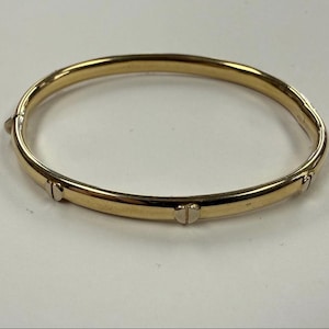 May include: A gold-coloured bangle bracelet. The bracelet is round with a hinged clasp and decorative accents. The metal has a polished finish, reflecting light. The bracelet is designed to be worn on the wrist.