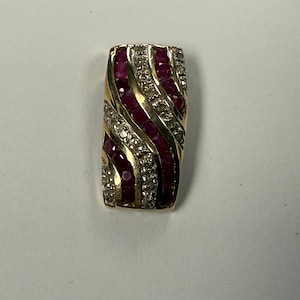 May include: A rectangular gold pendant featuring alternating bands of red gemstones and clear stones. The gemstones are arranged in a wavy pattern. This pendant is likely a piece of jewellery, with a striking design.