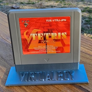 May include: A gray Virtual Boy game cartridge with a red and white label that says "TETRIS V・テトリス" and "VUE-VTRJ-JPN". The cartridge is sitting on a blue stand that says "VIRTUAL BOY".