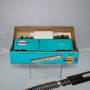 May include: A teal model train car from the New York Central System, labeled NYC 47062, sits inside its original box. The box is teal and brown with the Athearn Trains logo. Model railroad tracks are in the foreground.