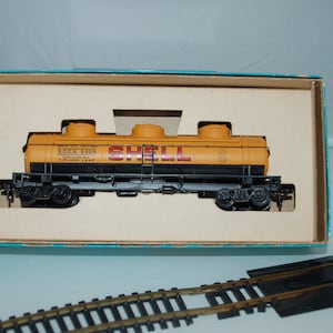 May include: A vintage model train car in a box. The yellow tank car has the Shell logo and text. The box is light blue and tan. The train car is black and yellow. The model train is on a light blue surface with a section of train tracks.