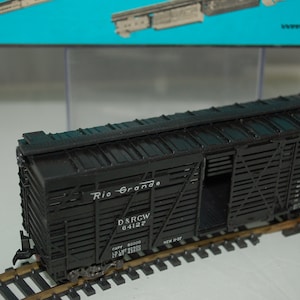 May include: A black model train boxcar with the words "Rio Grande" and other text on the side. The boxcar is on a model train track. The boxcar has an open door and is made of plastic.