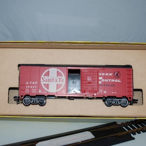 May include: A red Santa Fe model train boxcar with a white logo and text, inside a cardboard box. The boxcar has black wheels and the words "Hock Control" and "Smoother Ride" printed on the side. Model train tracks are in the foreground.