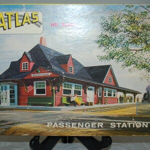 May include: A model train set box featuring a passenger station. The station is red with a black roof, and the sign reads "Rockville." The box has the "ATLAS" logo and the words "PASSENGER STATION."