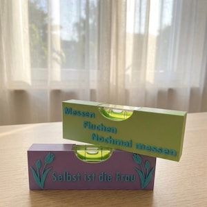 Op de afbeelding: Twee waterpassen, een lichtgroene en een paarse, liggen op een houten oppervlak. De groene waterpas heeft de blauwe tekst "Messen Fluchen Nochmal messen". De paarse waterpas heeft de blauwe tekst "Selbst ist die Frau".