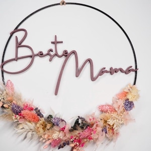 Puede incluir: Un adorno de pared circular con el texto "Best Mama" en escritura malva. El anillo de metal negro está adornado con un colorido arreglo floral de flores secas en tonos rosa, naranja y morado.