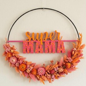 Könnte beinhalten: Ein kreisförmiger Wandschmuck mit den Worten "SUPER MAMA" in Orange und Rosa. Die Buchstaben sind vor einem rosa Hintergrund mit Leopardenmuster angeordnet. Der Kranz ist mit Trockenblumen in Orange- und Rosatönen verziert.