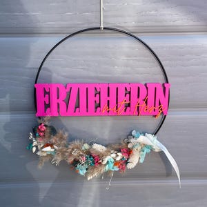 Könnte beinhalten: Ein dekorativer Kranz mit einem schwarzen Metallring, der das Wort "ERZIEHERIN" in leuchtendem Pink und "mit Herz" in Orange zeigt. Der Kranz ist mit getrockneten Blumen in Blau-, Rosa- und Beigetönen geschmückt.