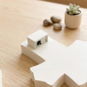 May include: A white, geometric-shaped object with a small, rectangular component on top, featuring a black button. The object is on a light-colored wooden surface, with a small succulent plant in a white pot and a few small stones in the background.