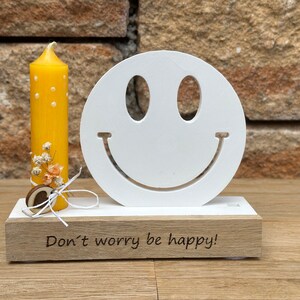Könnte beinhalten: Ein weißer Holz-Smiley-Kerzenhalter mit einer gelben Kerze und einem kleinen Blumenarrangement. Der Sockel des Halters trägt den Text "Keine Sorge, sei glücklich!" eingraviert.