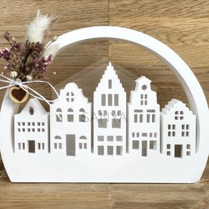 Könnte beinhalten: Weißer Holzausschnitt einer Häuserreihe mit einem halbrunden Torbogen darüber. Die Häuser haben Fenster und Türen. Ein Strauß getrockneter Blumen mit einem herzförmigen Holzzierstück befindet sich vor den Häusern.