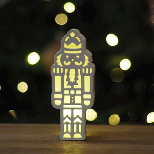 Könnte beinhalten: Weiße Holz-Nussknacker-Figur mit einem Licht im Inneren, das das Design beleuchtet. Der Nussknacker steht auf einer Holzoberfläche.