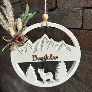 Könnte beinhalten: Weißer Holz-Kreis mit einer Bergszene, einem Reh und dem Wort "Bergliebe" in braunen Holzbuchstaben. Der Kreis hängt an einer Schnur mit einer Holzperle.