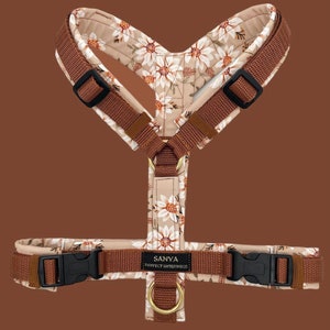 Op de afbeelding: Een bruin hondenharnas met een bloemenpatroon met witte madeliefjes en bruine stelen. Het harnas heeft een zwarte gesp en een gouden metalen ring. De tekst "SANYA PERFECT WINTERWOOL" is op het harnas gedrukt.