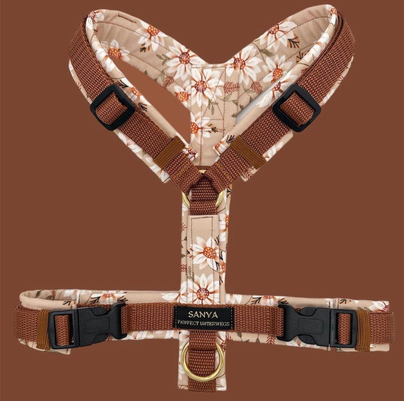 Op de afbeelding: Een bruin hondenharnas met een bloemenpatroon met witte madeliefjes en bruine stelen. Het harnas heeft een zwarte gesp en een gouden metalen ring. De tekst "SANYA PERFECT WINTERWOOL" is op het harnas gedrukt.