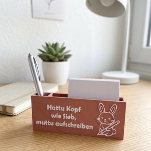 Op de afbeelding: Een terracotta bureau-organizer met drie compartimenten. De voorkant heeft witte tekst en een konijnenillustratie. Een zilveren pen en een stapel witte kaarten zitten erin. De tekst luidt "Hattu Kopf wie Sieb, muttu aufschreiben."