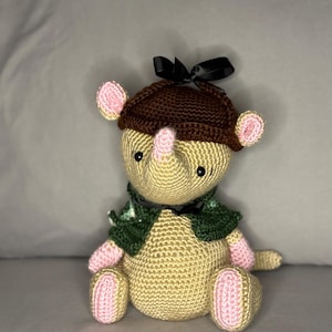 May include: A handmade crochet rhino toy in tan, pink, and brown. The rhino wears a brown hat with a black bow, a green scarf, and has pink accents on its ears, feet, and snout. The toy is sitting on a gray surface.