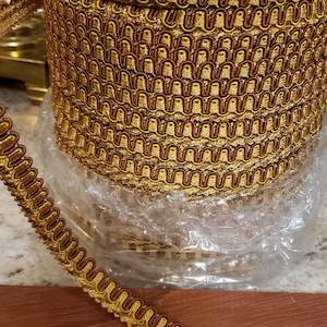 May include: A spool of decorative trim with a repeating pattern of gold and brown loops. The trim is made of a shiny material and is wrapped around a cardboard core. The trim is on a wooden surface.