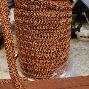 May include: A spool of decorative trim in a warm copper color. The trim features a detailed, looped design and is displayed on a wooden surface. The trim is ideal for sewing, crafting, and adding embellishments to various projects.