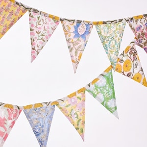 May include: A decorative bunting banner with floral patterns. The triangular flags display diverse flower designs in pink, yellow, blue, and green hues. The banner is strung together with a yellow trim.
