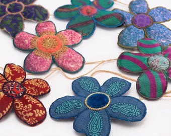 Puede incluir: Una guirnalda decorativa con múltiples flores de tela en varios colores y estampados, incluyendo morado, azul, verde, rosa y rojo. Cada flor está conectada por un cordel fino y los extremos de la guirnalda terminan en borlas de color beige.
