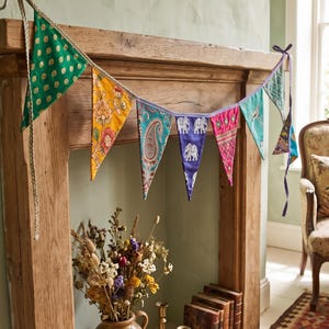 May include: A decorative bunting banner featuring a series of triangular fabric flags in various colours and patterns, including green with gold dots, yellow with floral prints, paisley, blue with white elephants, pink, and teal with bird motifs.